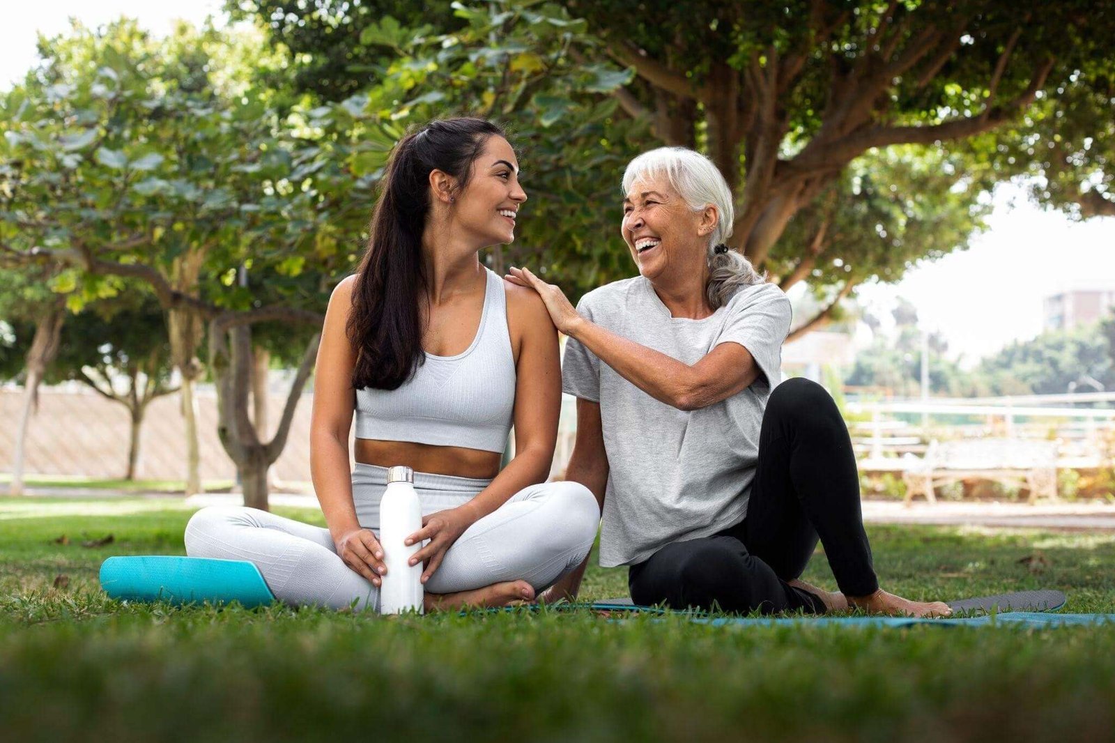 Duas mulheres com roupa de ginástica sentadas na grama de um parque pela manhã conversando e rindo.