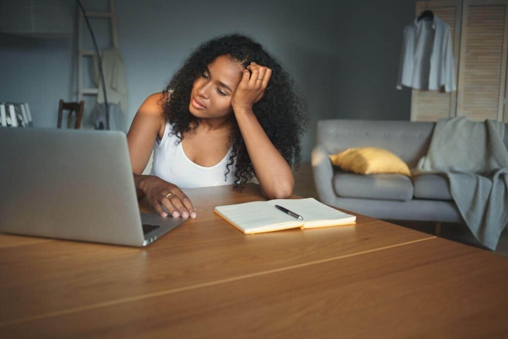 Mulher sentada à mesa de trabalho com a mão no rosto, olhando para um notebook com expressão de desânimo e bloqueio criativo. O cenário representa a estafa cognitiva e a necessidade de nutrientes e vitaminas para cansaço mental.